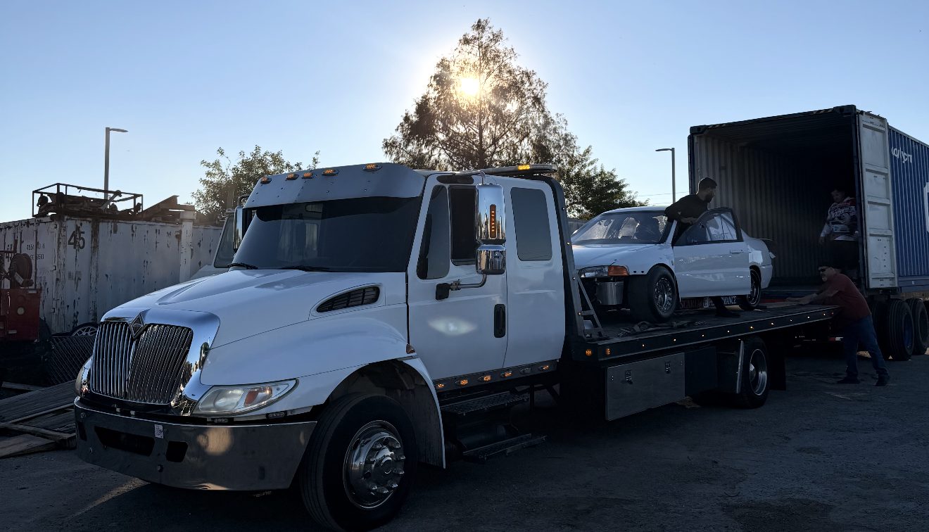 A flatbed tow truck loading a car at a recovery yard with the sun low on the horizon